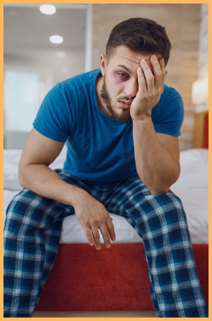 A candid image of a tired man sitting on the edge of a bed, dressed in a blue t-shirt and plaid pajama pants. He rests his head in one hand, his weary expression reflecting exhaustion or deep thought, capturing the struggle of inner dialogue or the weight of a long, draining day